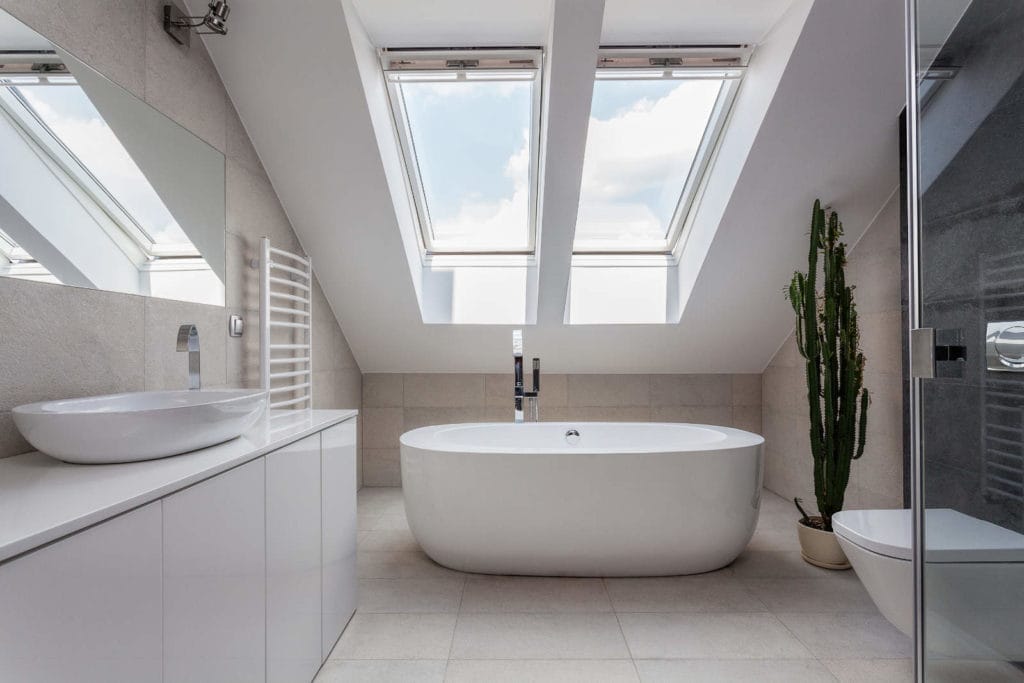 Modernes Badezimmer mit freistehender ovaler Badewanne unter Oberlichtern. Es verfügt über ein weißes Waschbecken auf einer Ablage, einen hohen Kaktus und einen Handtuchhalter. Boden und Wände sind mit grauen Fliesen ausgelegt, rechts befindet sich eine Duschkabine aus Glas.