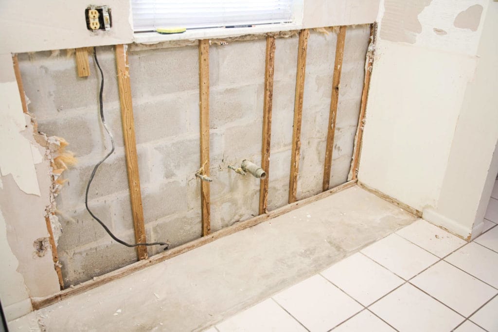 Eine Küchenwand wird renoviert, mit freiliegenden Ständern und Betonblöcken. Links sind Stromleitungen sichtbar. Die alte Trockenbauwand wurde teilweise entfernt, in der Mitte sind die Sanitärrohre freigelegt. Der Boden ist weiß gefliest.