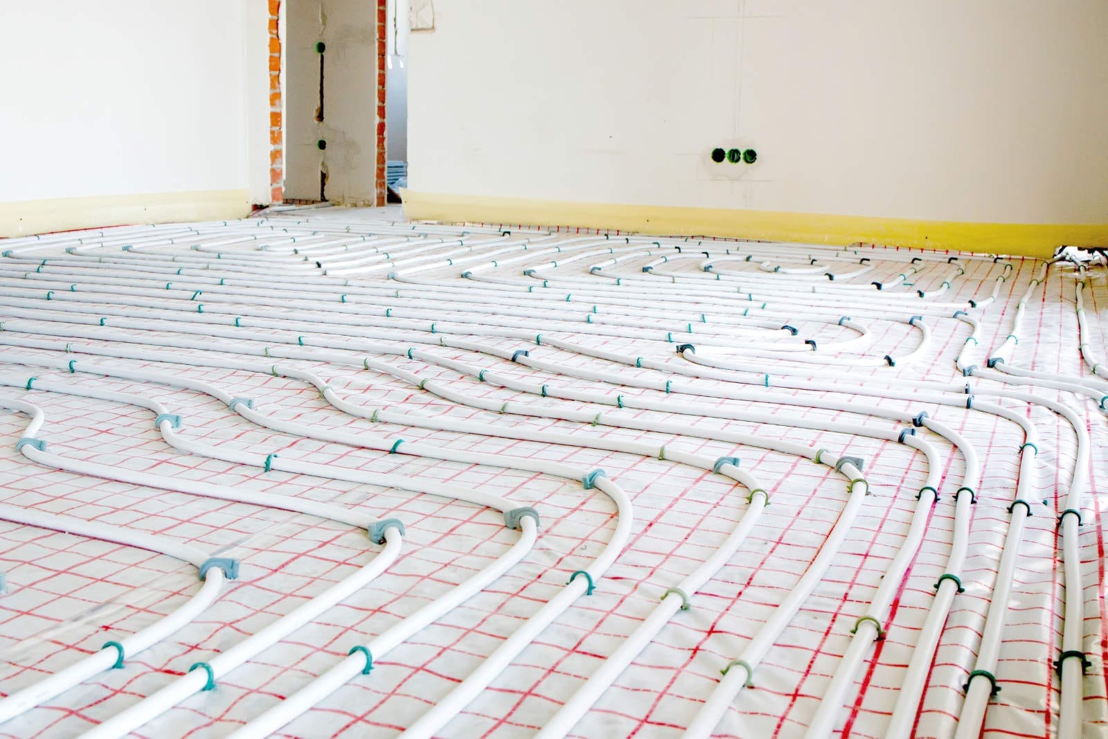 Bild eines im Bau befindlichen Raumes mit Trockenbauelementen. Auf dem Boden ist ein Netz aus weißen Rohren verlegt, möglicherweise für eine Fußbodenheizung, auf einer gitterförmigen Kunststofffolie. Die Wände sind noch nicht fertiggestellt, sichtbare Steckdosen warten auf Malerarbeiten.
